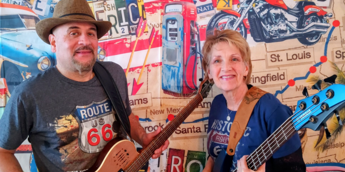 The Victor Samalot Duo smiling and holding guitars while standing in front of a decorative Route 66 wallpaper background.