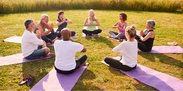 A sunny and grassy setting with eight adults sitting on mats arranged in a circle, hands in prayer position.