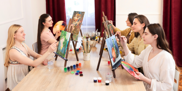 A group of adults sitting at a table with painting supplies working on individual paint canvases on easles.