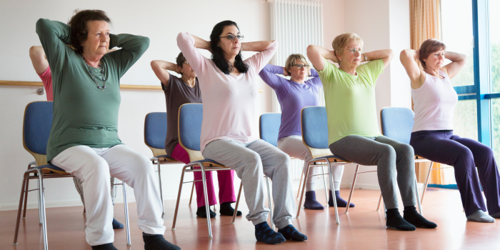 Small group of adults in a yoga position with arms raised, hands clasped behind head while sitting in chairs.