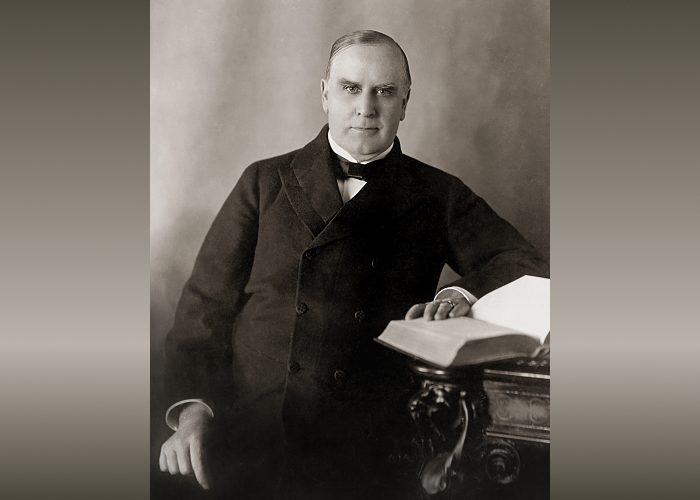​​President William McKinley sitting at desk with hand resting on an open book.