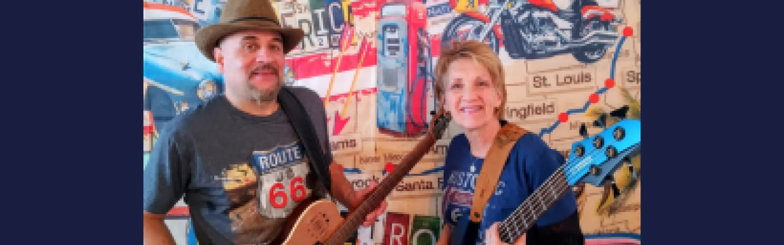 The Victor Samalot Duo smiling and holding guitars while standing in front of a decorative Route 66 wallpaper background.