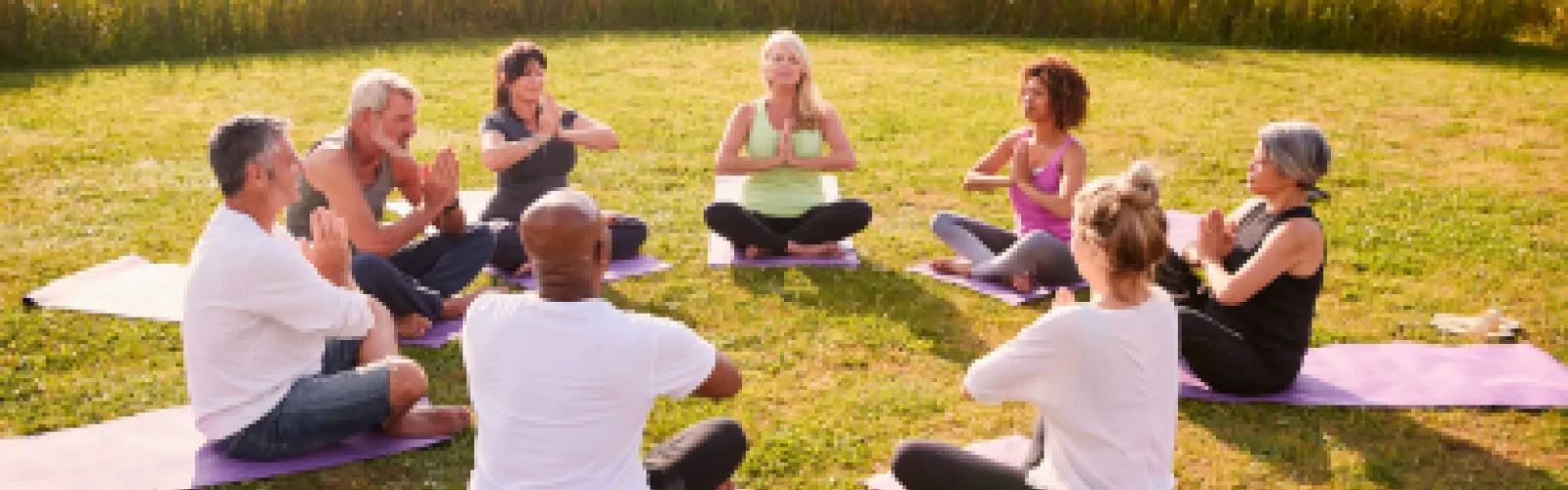 A sunny and grassy setting with eight adults sitting on mats arranged in a circle, hands in prayer position.